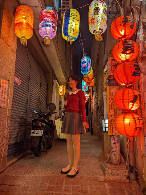 A full-body photograph of the author, taken during nighttime. She is dressed in a skirt, blouse, and platform shoes. Her straight black hair falls just above her shoulder. She smiles and stands in front of an alleyway decorated with paper lanterns.